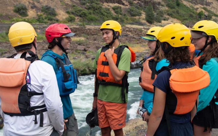 a group of outward bound students wearing helmets and life jackets talk to each other while standing above a river